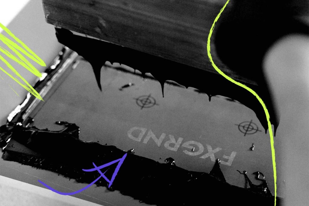 Nahaufnahme eines Siebdruckrahmens mit schwarzer, verlaufender Druckfarbe; das spiegelverkehrte Logo „FXGRND“ ist auf dem Sieb sichtbar, daneben handgezeichnete Markierungen und Pfeile